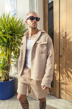 Güneş gözlüklü yakışıklı genç adam moda kıyafetleri içinde poz veriyor.