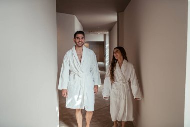 Bornozlu genç çift otelde yürürken dinleniyor ve gülümsüyor.