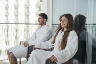 Otel odasında bornozun yanında oturan mutlu genç çift.