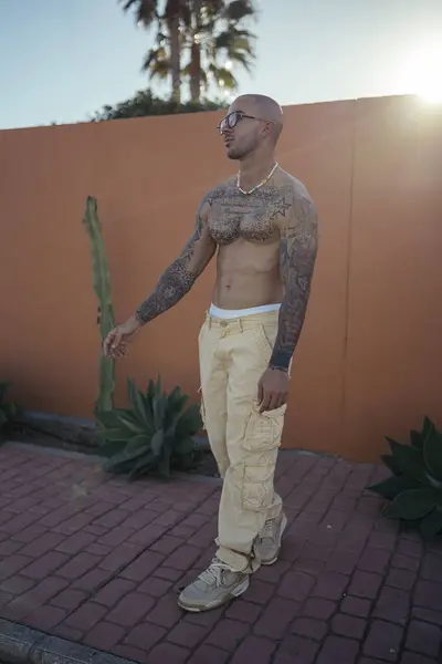 Şehirde poz veren güneş gözlüğü ve dövme takan yakışıklı genç erkek modelin portresi..