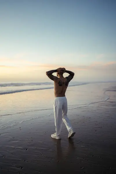 Yakışıklı bir adam kumsalda beyaz gömlekli duruyor ve gün batımını seyrediyor.