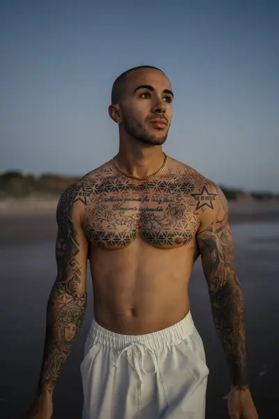 Kumsalda vücudunda dövmeleri olan yakışıklı, üstsüz bir adam.