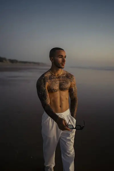 Ellerini kumda tutan genç yakışıklı adamın portresi. Kumsalda duran beyaz tişörtlü ve pantolonlu yakışıklı bir erkek..