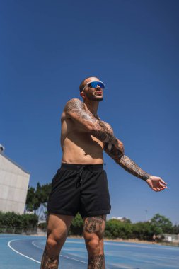 Plajda mavi gömlekli, şortlu güneş gözlüğü takan dövmeli genç bir adam. Adam yolda koşuyor.