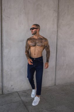 Şehirde poz veren güneş gözlüklü ve dövmeli yakışıklı bir genç.