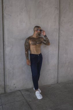 Güneş gözlüklü ve kot pantolonlu yakışıklı adam gri duvar arkasında duruyor.