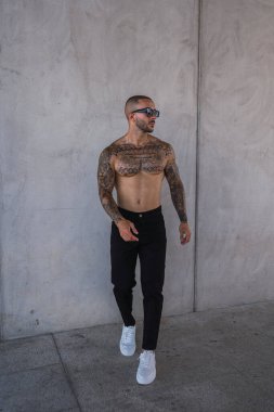 Sakallı ve güneş gözlüklü genç yakışıklı bir adam gri arka planı olan beyaz bir duvarın karşısında duruyor..