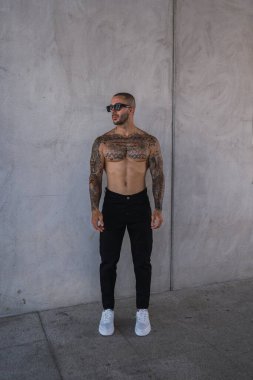 Beyaz tişörtlü ve siyah pantolonlu bir adam sokakta duruyor. Dövmeli ve güneş gözlüklü beyaz tişörtlü adam..