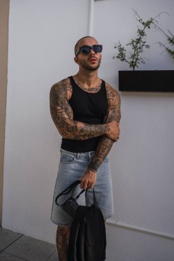 Omzunda dövmeleri olan, şık siyah tişört ve güneş gözlüğü takan yakışıklı genç bir adamın modern şehir bölgesinde duran portresi.