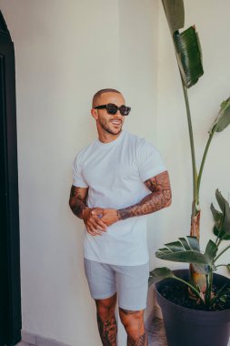 Güneş gözlüklü ve tişörtlü genç Afro-Amerikan model iç mekânda poz veriyor.