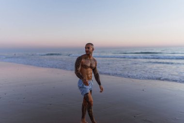 Genç, üstsüz adam kollarını göğsüne dayayıp kameraya bakıyor.