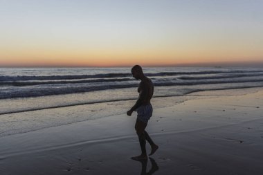 Gün batımında sahilde sörf tahtası olan bir adamın silueti.