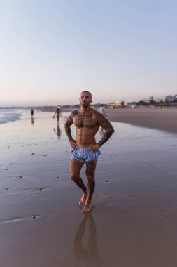 Üstsüz bir adam sahilde dikilip kameraya bakıyor.