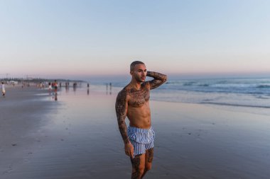 Gün doğumunda plajda dikilen dövmeli genç üstsüz adam.