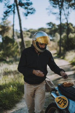 Kasklı ve ceketli, güneş gözlüklü bir erkek motosikletçi yolun kenarında motosikletli duruyor.