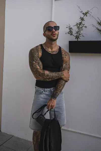 Omzunda dövmeleri olan, şık siyah tişört ve güneş gözlüğü takan yakışıklı genç bir adamın modern şehir bölgesinde duran portresi.