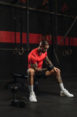 Genç, kaslı bir erkek sporcu bankta oturuyor ve elinde bir halter tutuyor..
