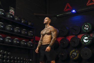 Siyah tişörtlü ve siyah şortlu genç yakışıklı spor salonunda duruyor. Kaslı adam başka tarafa bakıyor ve gülümsüyor.