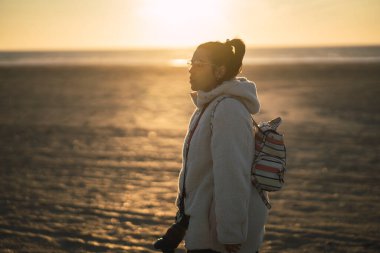 Genç bir kadın gün batımında sahilde yürüyor.