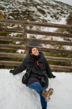 Kırmızı atkısı ve beyaz çizmeleri olan mutlu bir kız kış ormanında karda oturuyor.