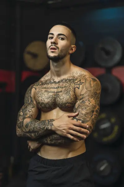 Siyah bir duvarın önünde poz veren dövmeli, kaslı, genç, üstsüz bir adam..