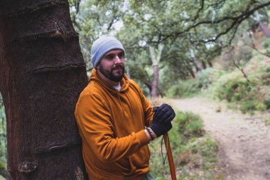 Sakallı, bıyıklı, turuncu ceketli, şapkalı, sonbahar ormanında sırt çantalı bir adam kameraya bakıyor.