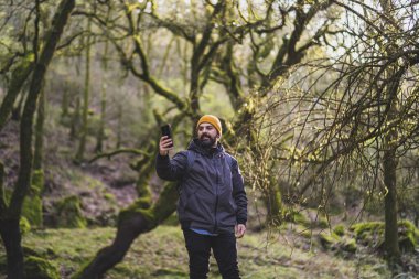 Siyah ceketli sakallı ve arka planda cep telefonu olan bir adam..