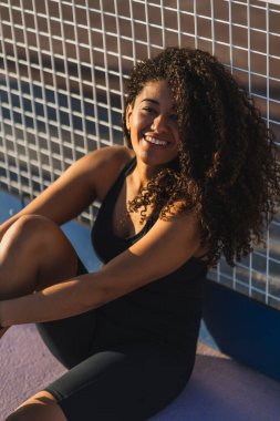 Genç siyah kadın bankta oturuyor ve kameraya bakıyor.