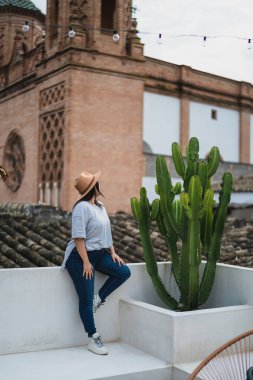 Parkta bir kaktüsün üzerinde oturan güneş gözlüklü ve şapkalı kadın.