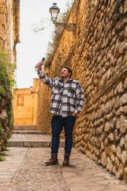 Şapkalı ve gömlekli genç adam cep telefonlu taş bir duvarda oturuyor..