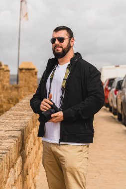 Sırt çantalı, sakallı, beyaz tişörtlü ve arka planda sakallı yakışıklı bir genç adam.