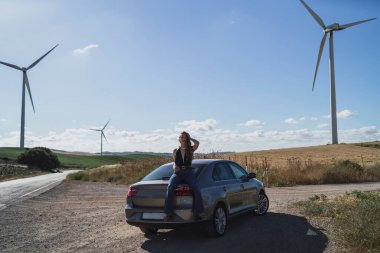 Bir kadın arabanın yanında duruyor ve yolda rüzgar türbini tutuyor.