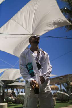 Güneş gözlüklü ve şapkalı mutlu adam arka planda havuzda poz veriyor.