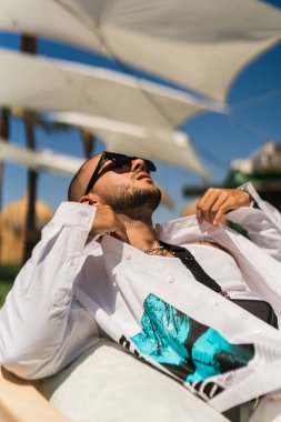 Güneş gözlüklü ve beyaz gömlekli genç adam plajda yatıyor.