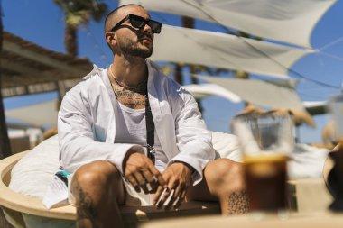 Beyaz gömlekli genç bir adamın portresi bir sandalyede oturuyor ve sahilde kahve içiyor, yaz tatilinde.