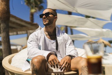 Güneş gözlüklü ve siyah takım elbiseli genç adam plaj koltuğunda oturuyor.