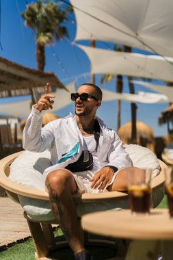 Güneş gözlüklü genç adam sahilde kokteyl içiyor.