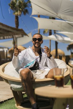 Güneş gözlüklü genç adam dinlenme odasında dinleniyor ve yaz tatilinde eğlenirken kokteyl içiyor.