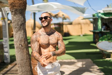 Kafasında dövmeleri olan ve kameraya gülümseyen güneş gözlüklü genç bir adam.