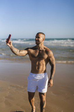 Adam akıllı telefonuyla kumlu bir sahilde selfie çekiyor.