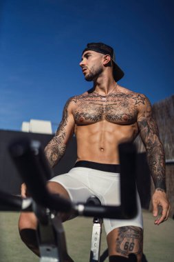 Bisikletli üstsüz, kaslı bir adam güneşli bir günde dışarıda dinlenirken yüzünü çeviriyor.