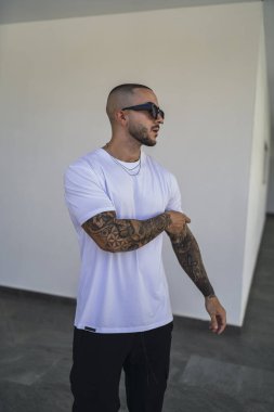Genç, yakışıklı, güneş gözlüklü, dövmeli, beyaz tişörtlü bir adam..