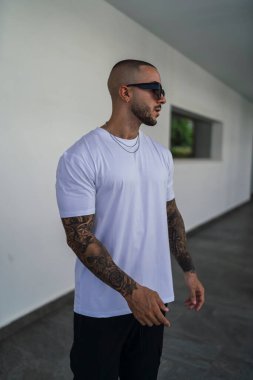 Metal bir yapısı olan beton bir duvarın arka planında siyah güneş gözlüğü olan beyaz tişörtlü genç bir adam..