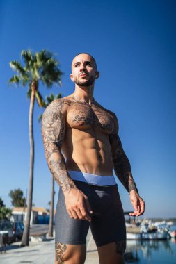 Yazın sahilde poz veren dövmeli yakışıklı, güçlü, kaslı genç bir adam.