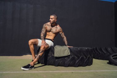 Duvara yaslanırken spor topunun üzerinde lastik tutan üstsüz bir adam.