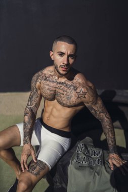 Üstsüz, kaslı, dövmeli, gri arka planda poz veren adam.