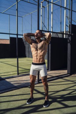 Parkta çitin üzerinde beyzbol topuyla poz veren üstsüz kaslı genç adam.