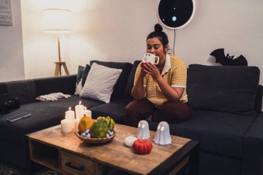 Genç siyah kadın koltukta oturuyor ve elleriyle çay içiyor.