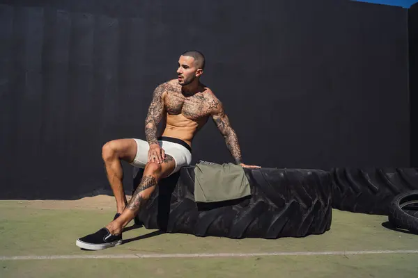 Duvara yaslanırken spor topunun üzerinde lastik tutan üstsüz bir adam.