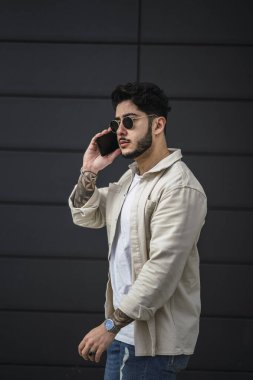 Güneş gözlüklü ve siyah ceketli genç adam duvarda poz veriyor.